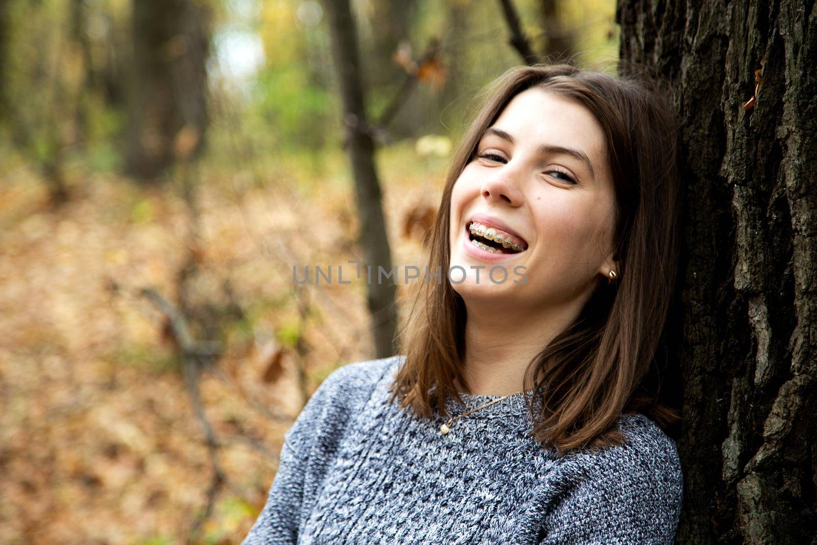 Young beautiful girl with braces on her teeth in a gray sweater sits in the autumn forest near a large tree and smiles sweetly.