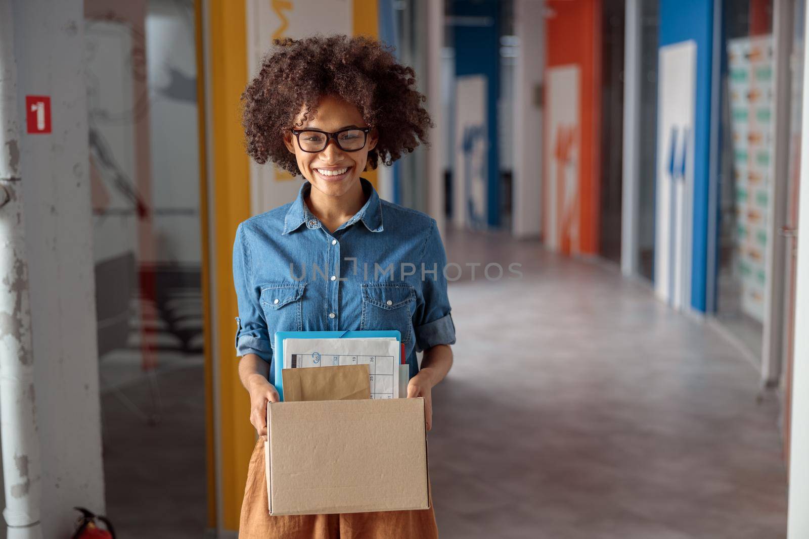 Happy Afro American woman in glasses standing in the hallway while holding paper box, copy space