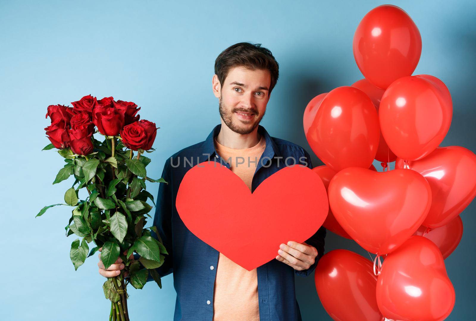 Cute guy waiting for girlfriend with valentines day presents, holding bouquet of roses and red heart, smiling at camera, standing over blue background by Benzoix