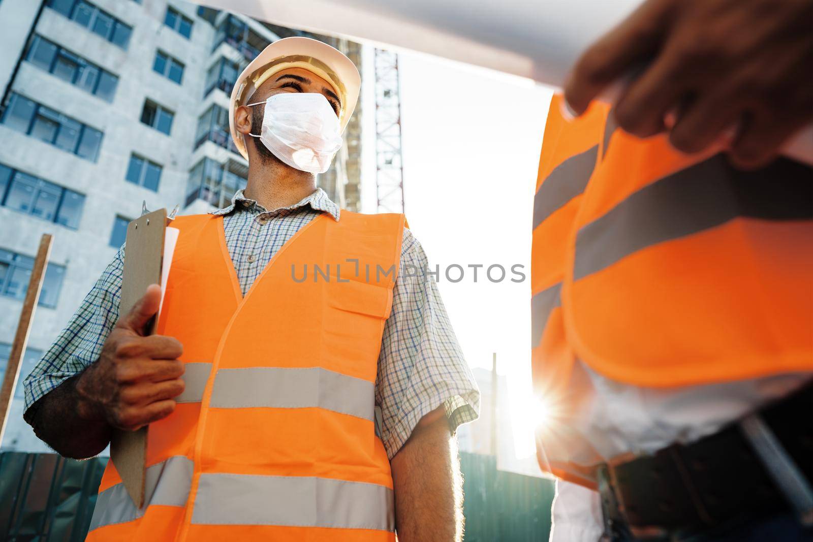 Two men in workwear and medical masks working with blueprints on object, close up