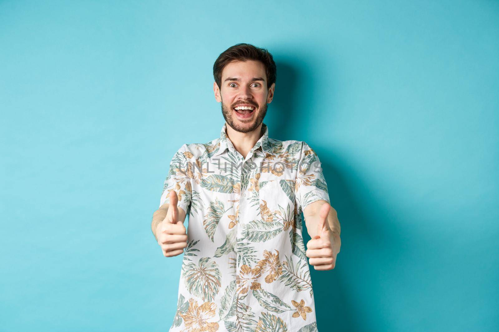 Excited smiling guy in hawaiian shirt show thumbs up, looking amazed, checking out cool promo, standing on blue background.