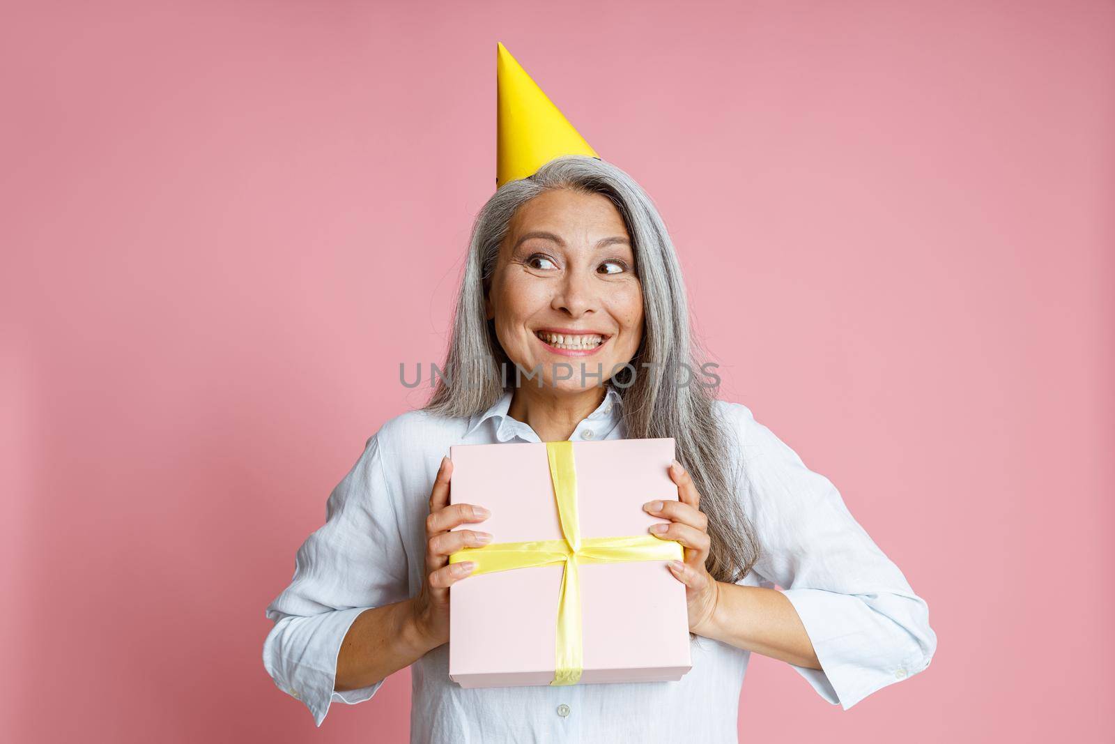 Emotional mature Asian woman with loose grey hair and yellow party hat holds present posing on pink background in studio
