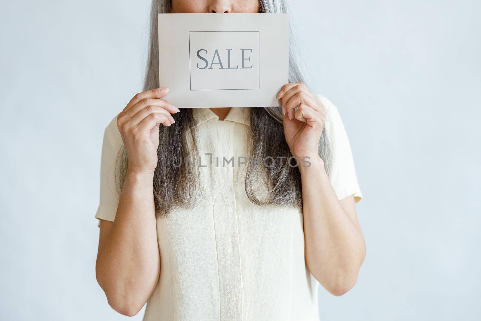 Mature woman with long grey hair holds Sale sign near face standing on light background in studio closeup