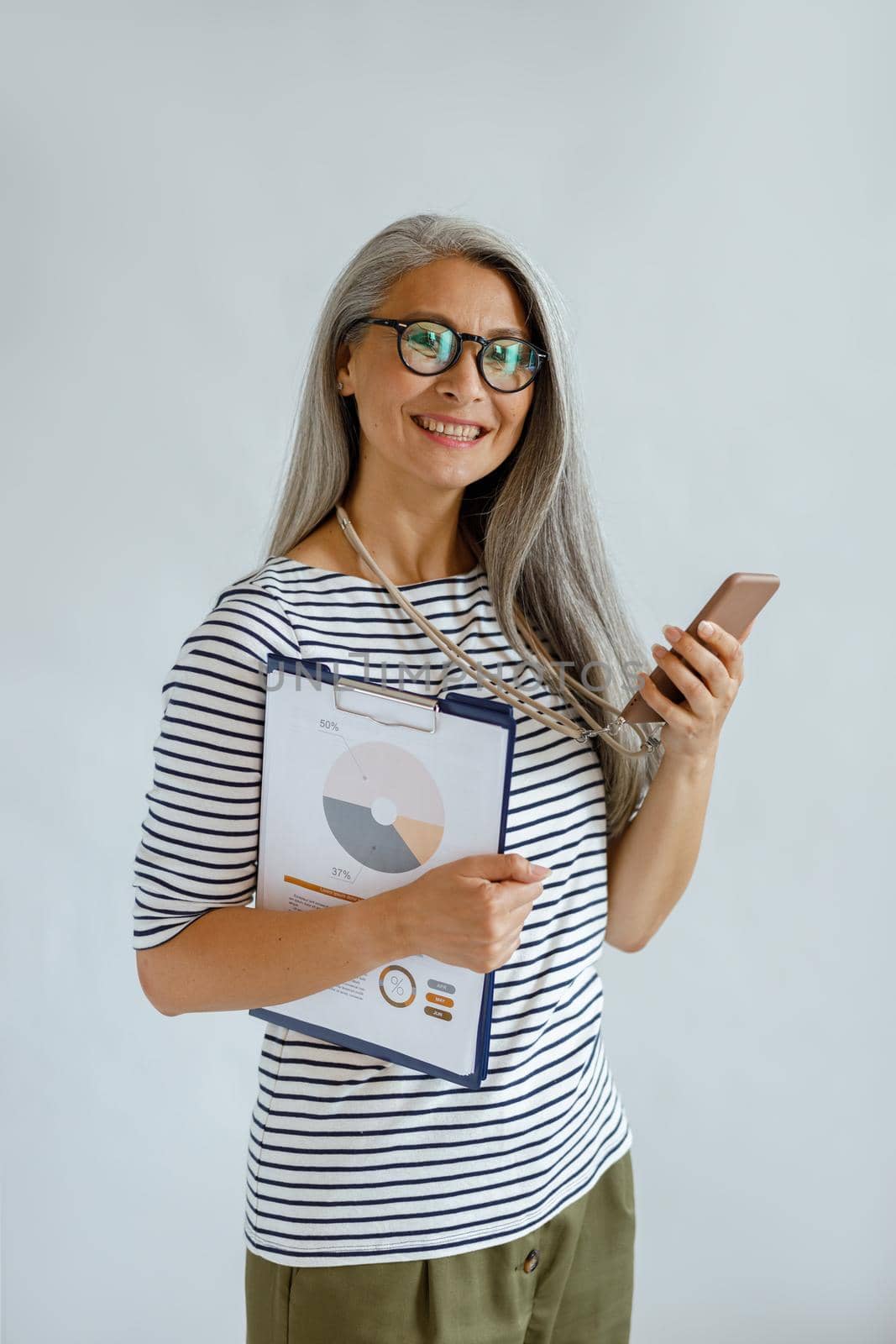 Smiling silver haired Asian lady with glasses holds colorful diagrams and smart phone standing on light grey background in studio