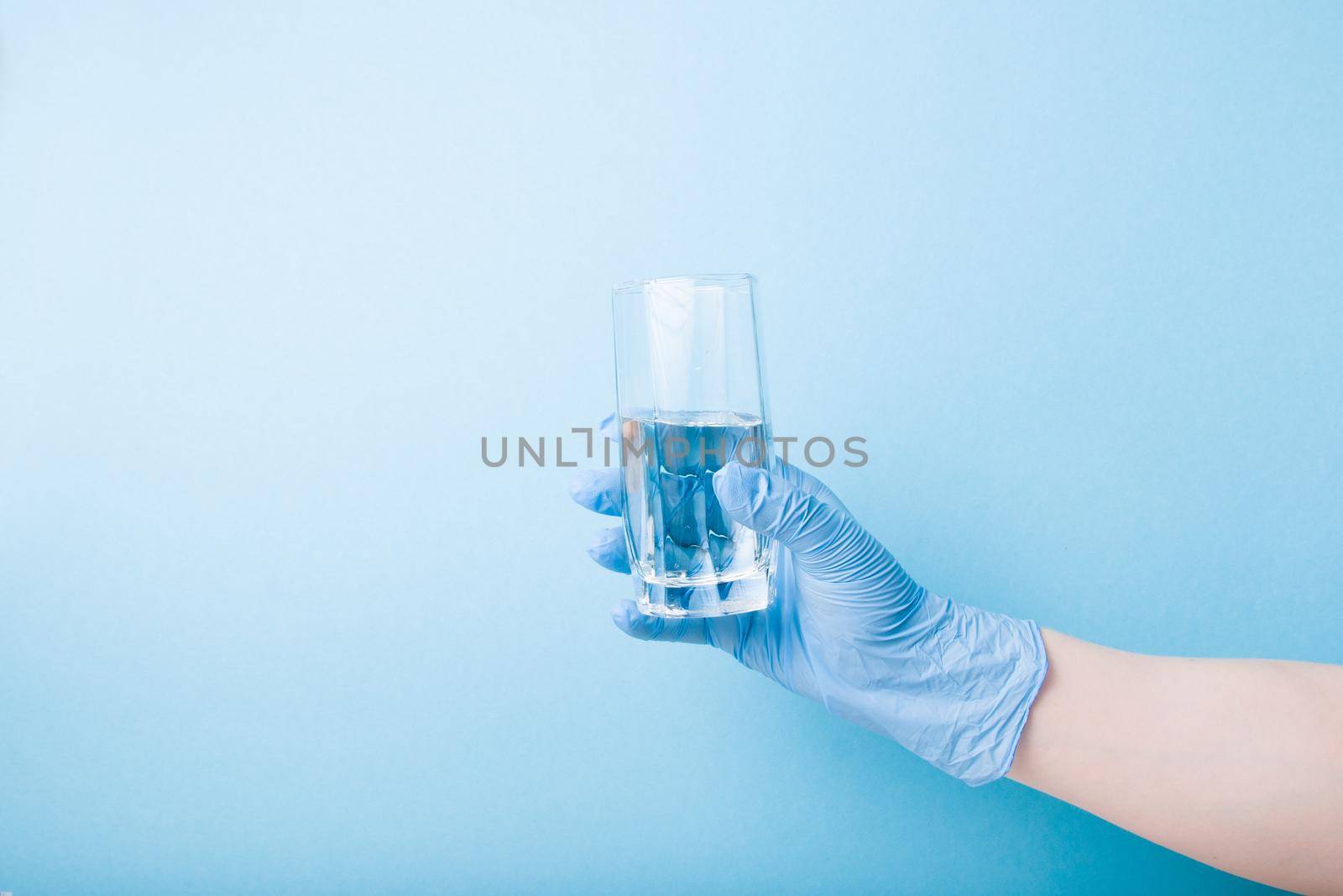 hand in blue disposable medical glove holds a glass glass with water, blue background copy space