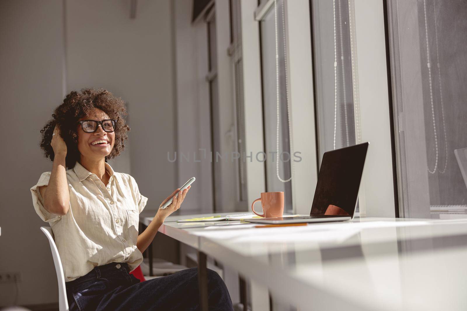 Happy young lady in headphones and holding mobile phone while sitting at her workplace