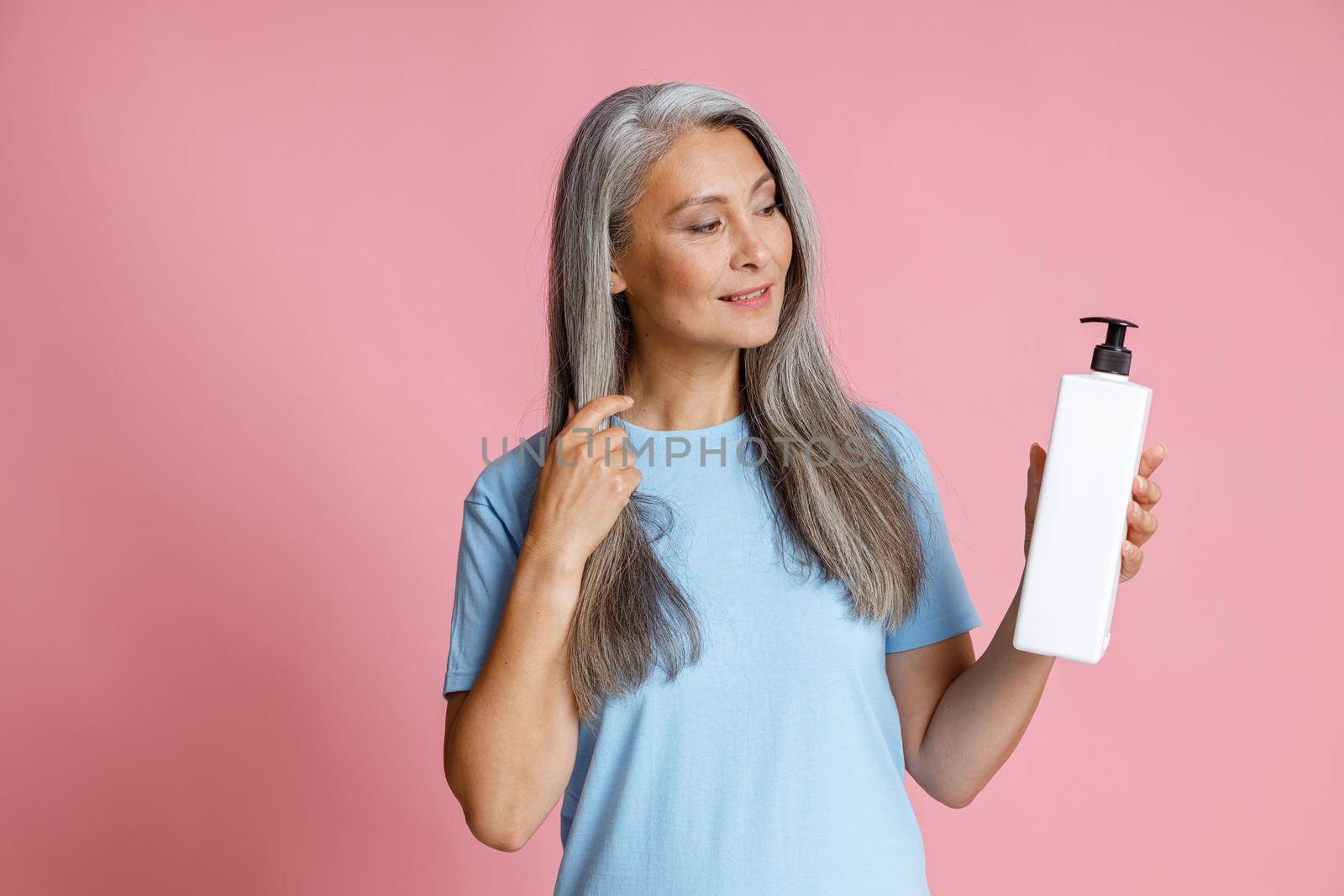 Beautiful Asian woman with loose silver hair holds blank dispenser bottle of cosmetic products on pink background in studio. Mature beauty lifestyle