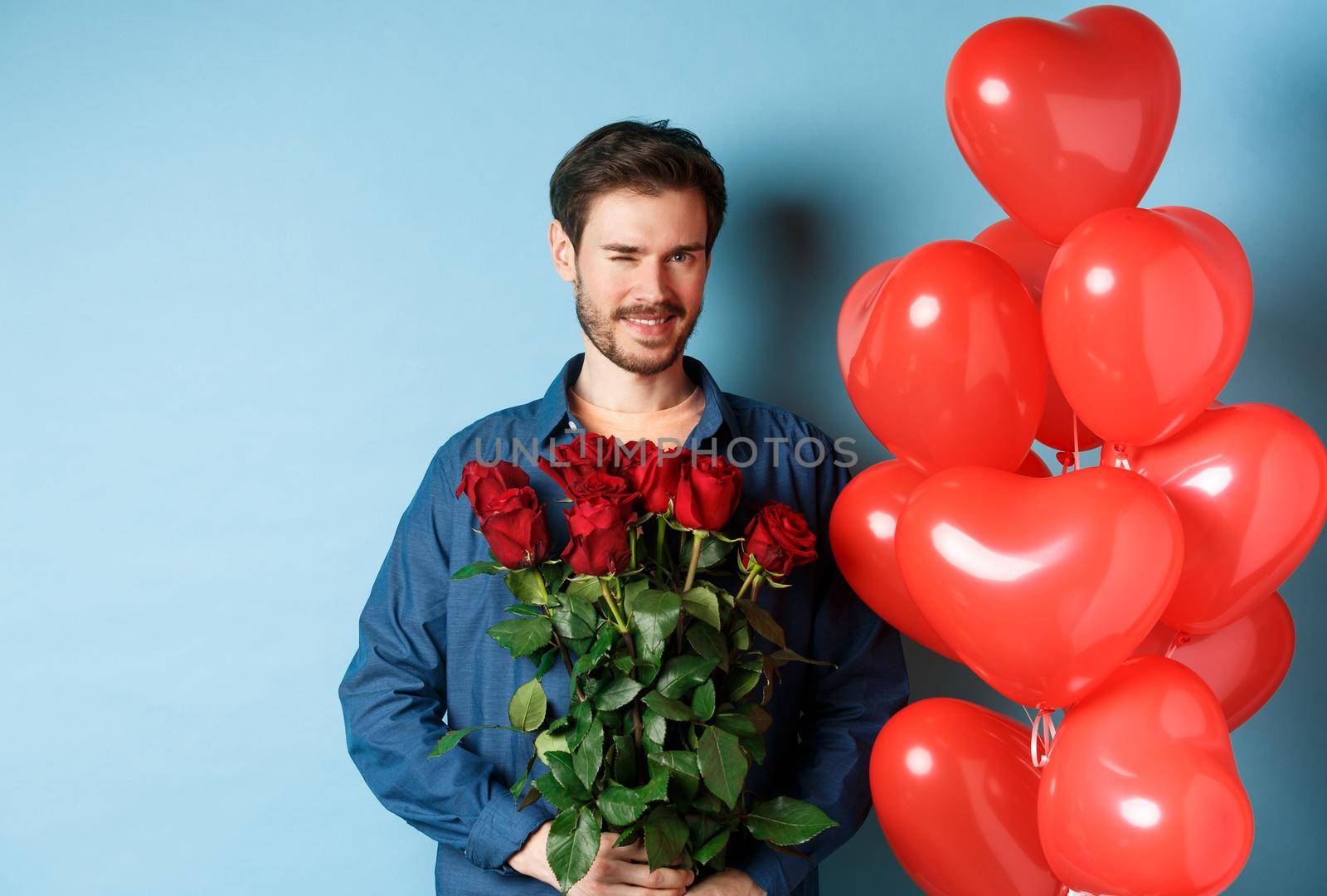 Romantic boyfriend winking and smiling, holding bouquet of flowers on Valentines day, standing near heart balloons for lover, blue background.