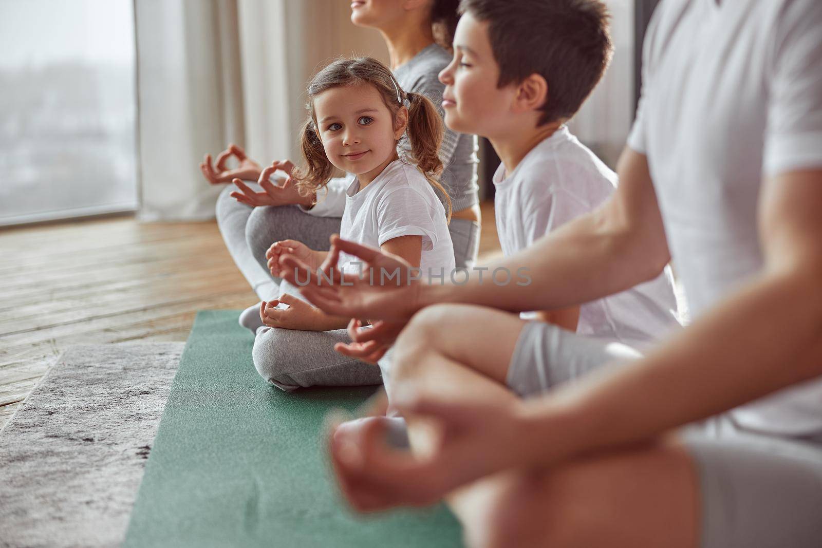 Cheerful daughter is sitting in lotus position near older brother and calm parents at home