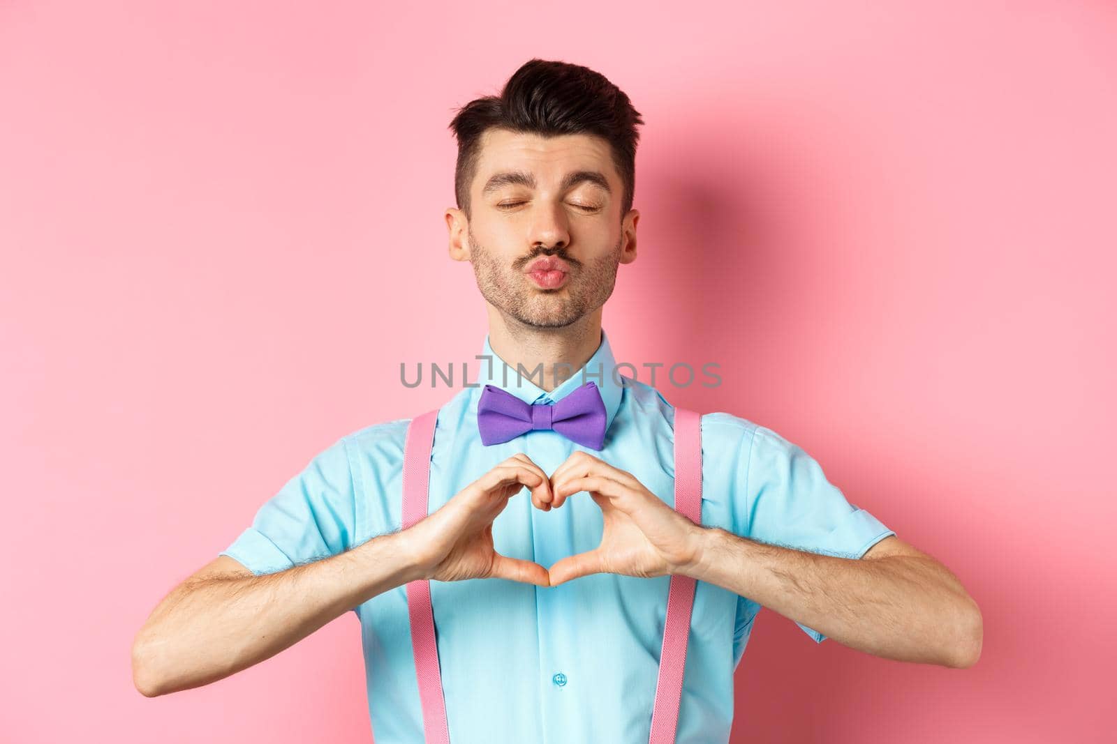 Happy Valentines day. Romantic boyfriend close eyes for kiss and showing heart, having date with lover, standing over pink background.