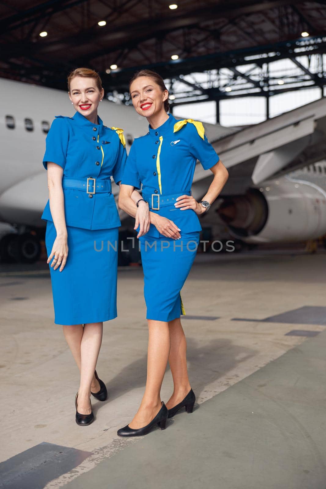 Full length shot of two air stewardesses in stylish blue uniform smiling at camera, standing together in front of passenger aircraft in hangar at the airport by Yaroslav_astakhov