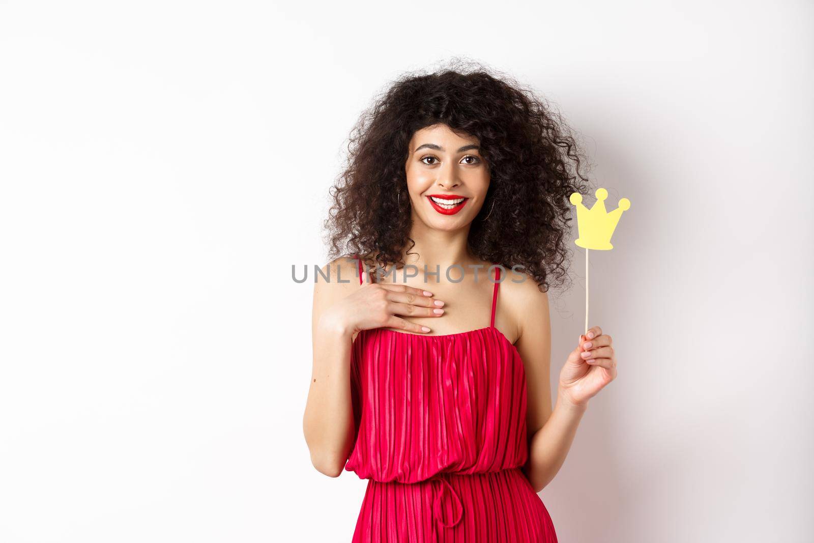 Confident pretty lady in red dress, holding queen crown on stick and looking excited, standing on white background.
