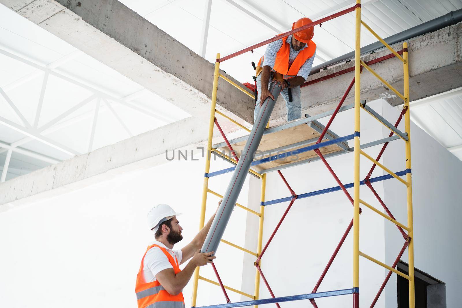 Two men builder in uniform drag the pipe on scaffolding at construction site by Fabrikasimf