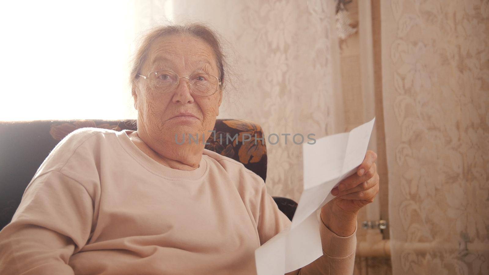 An elderly woman sitting in a chair and reading something, looking surprised not in a good way. Looking in the camera. Close up