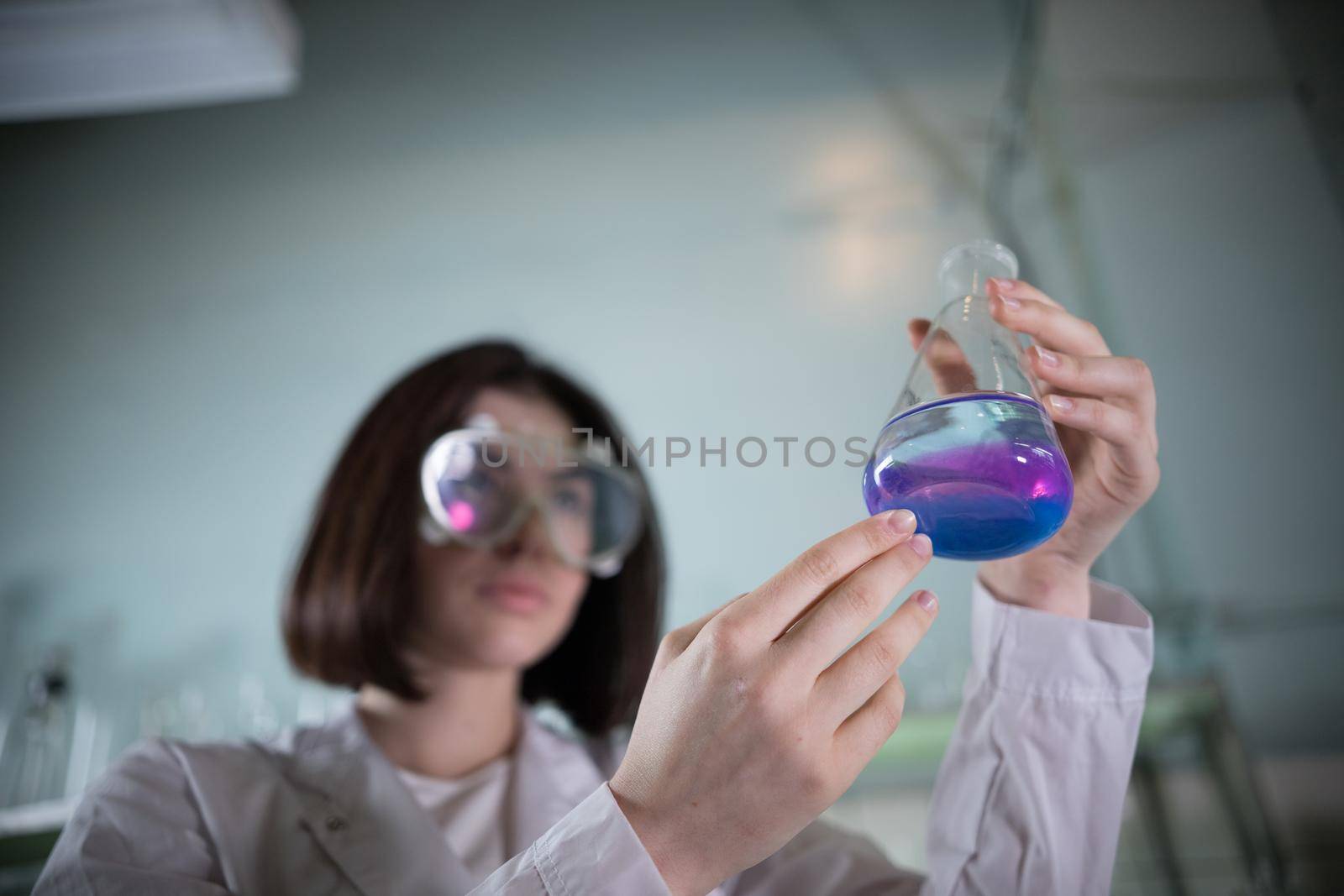 Chemical laboratory. Young woman holding a flask with blue and purple exfoliating liquid in it. Flask in focus
