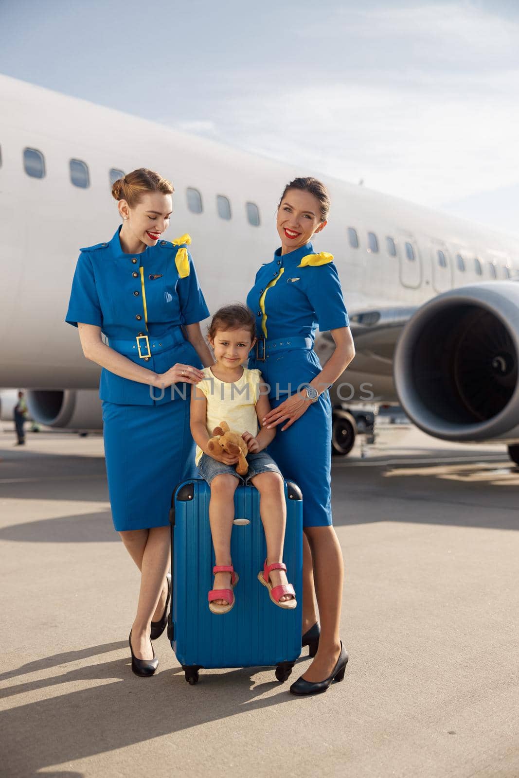 Full length shot of smiling beautiful stewardesses standing near cute little girl sitting on suitcase in front of big airplane and smiling at camera by Yaroslav_astakhov