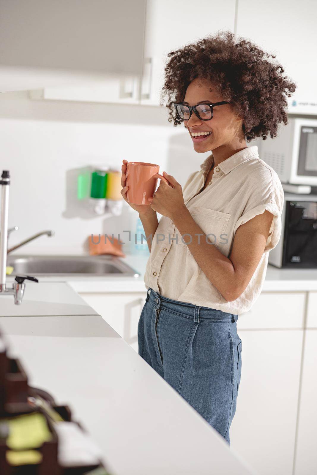 Happy multiethnic woman in glasses standing and holding cup of coffee on coworking kitchen