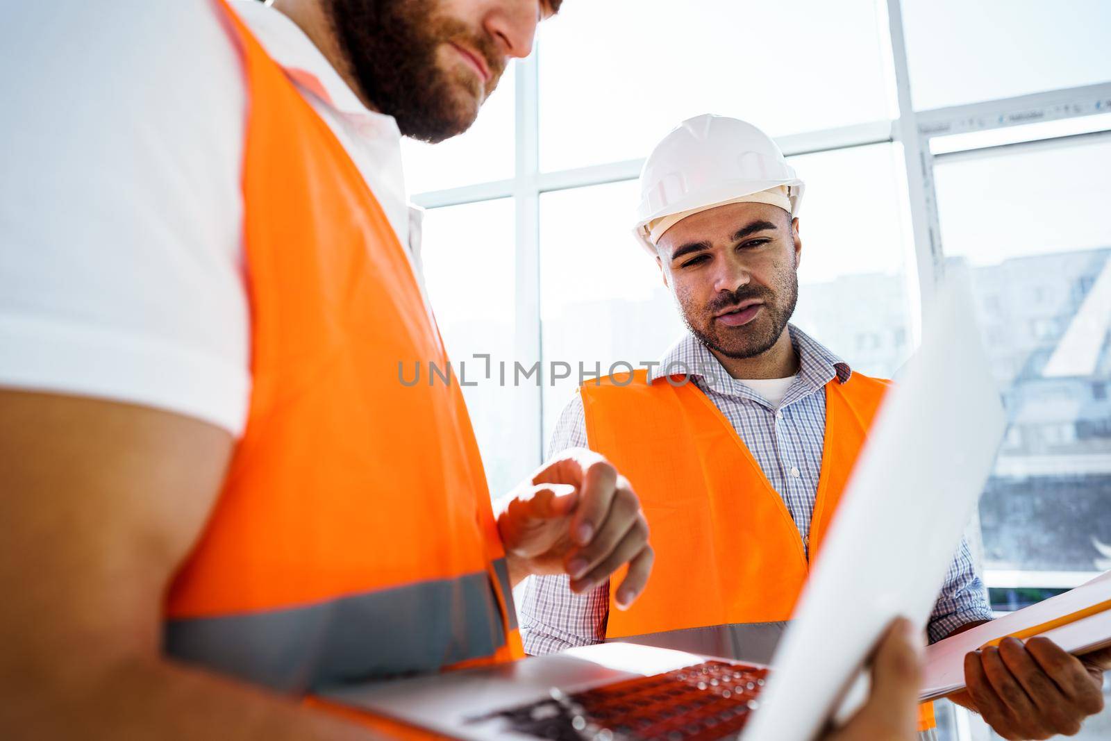 Two specialists supervisors in hardhats using laptop at construction site for work by Fabrikasimf