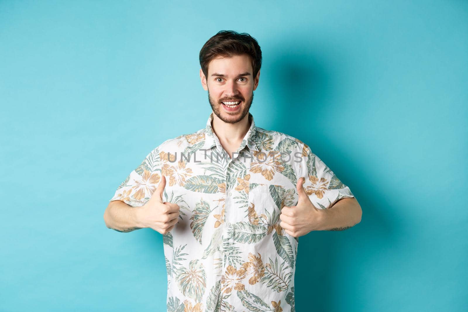 Handsome tourist showing thumbs up and say yes, praising good tourism agency, wearing hawaiian shirt, standing on blue background.