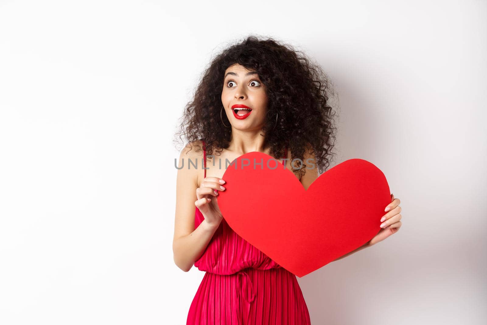 Valentines day. Young woman looking surprised left, seeing something amazing on lovers day, found true love, holding big red heart on chest, white background by Benzoix