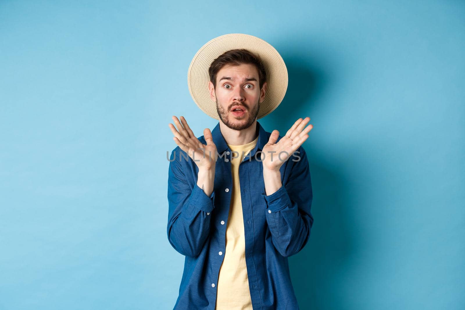 Image of shocked tourist in straw hat panicking, raising hands up and stare startled at camera, standing on blue background.