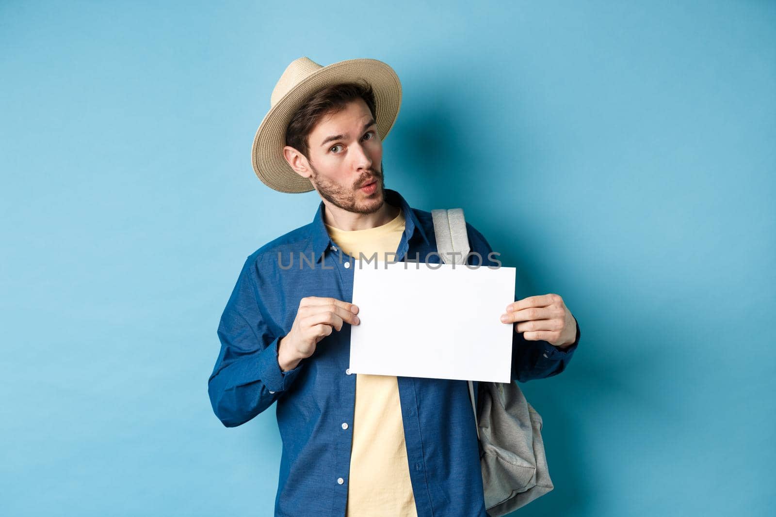 Excited tourist in straw hat, hitchhiking, showing blank piece of paper and looking amused, standing on blue background. Copy space