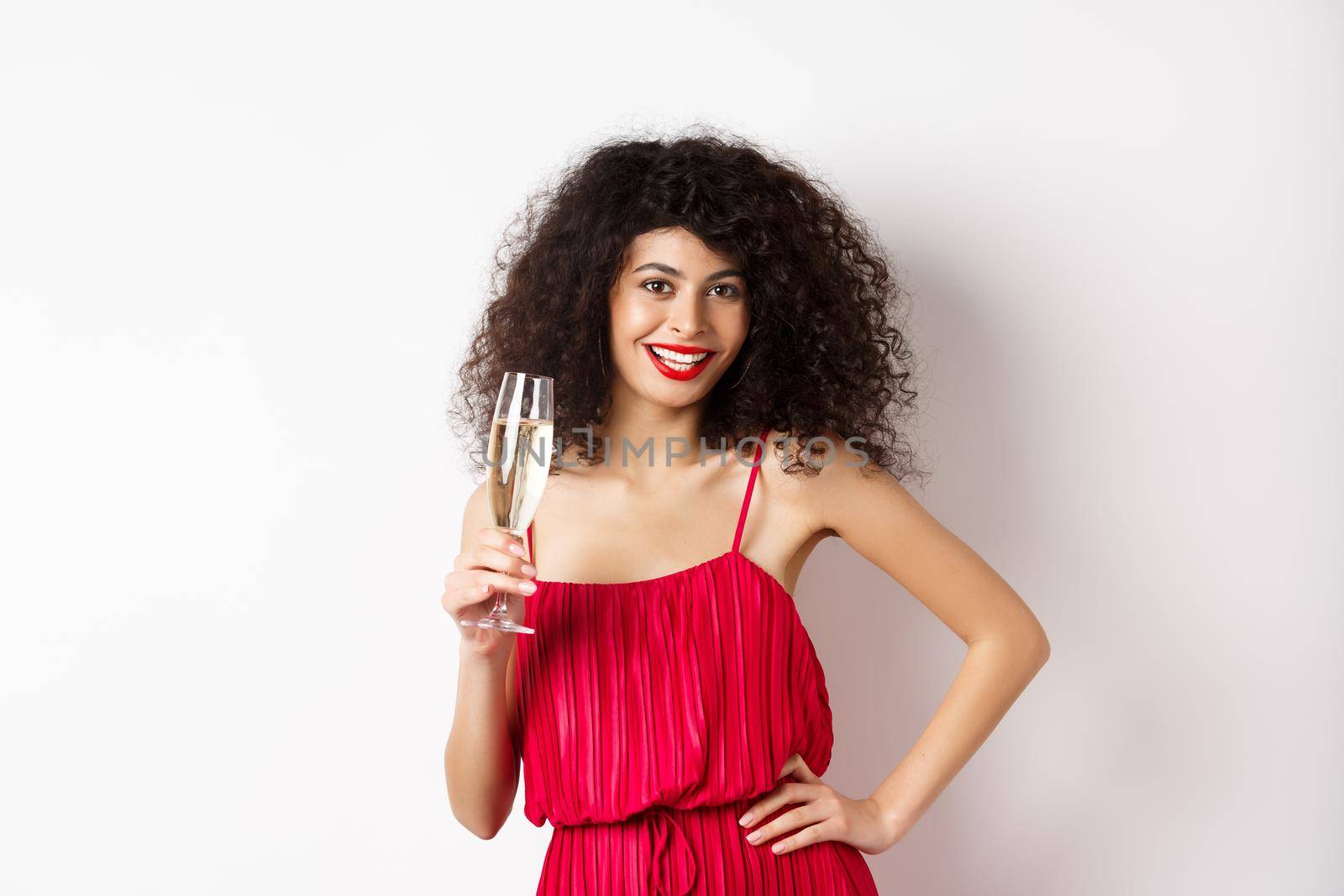 Sassy young woman in elegant red dress, posing with glass of champagne on white background, having romantic date on valentines day.