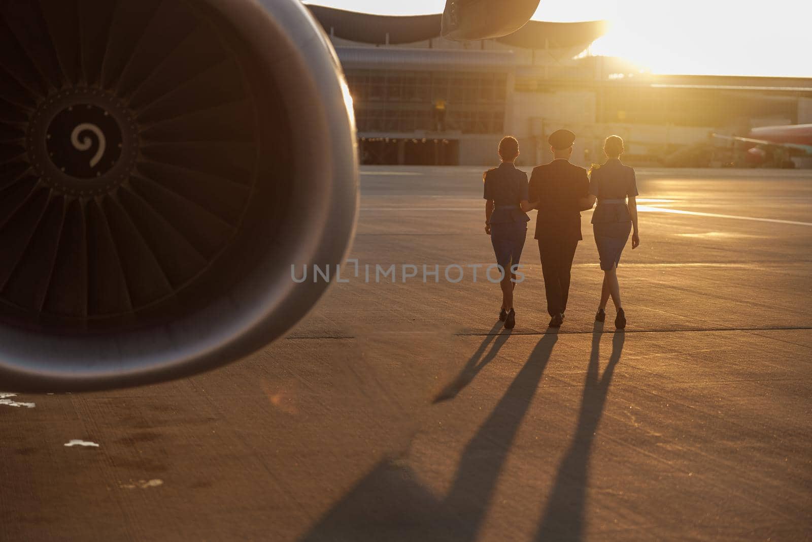 Full length shot of professional pilot walking together with two female flight attendants in blue uniform in terminal at sunset. Aircraft, aircrew concept. Back view