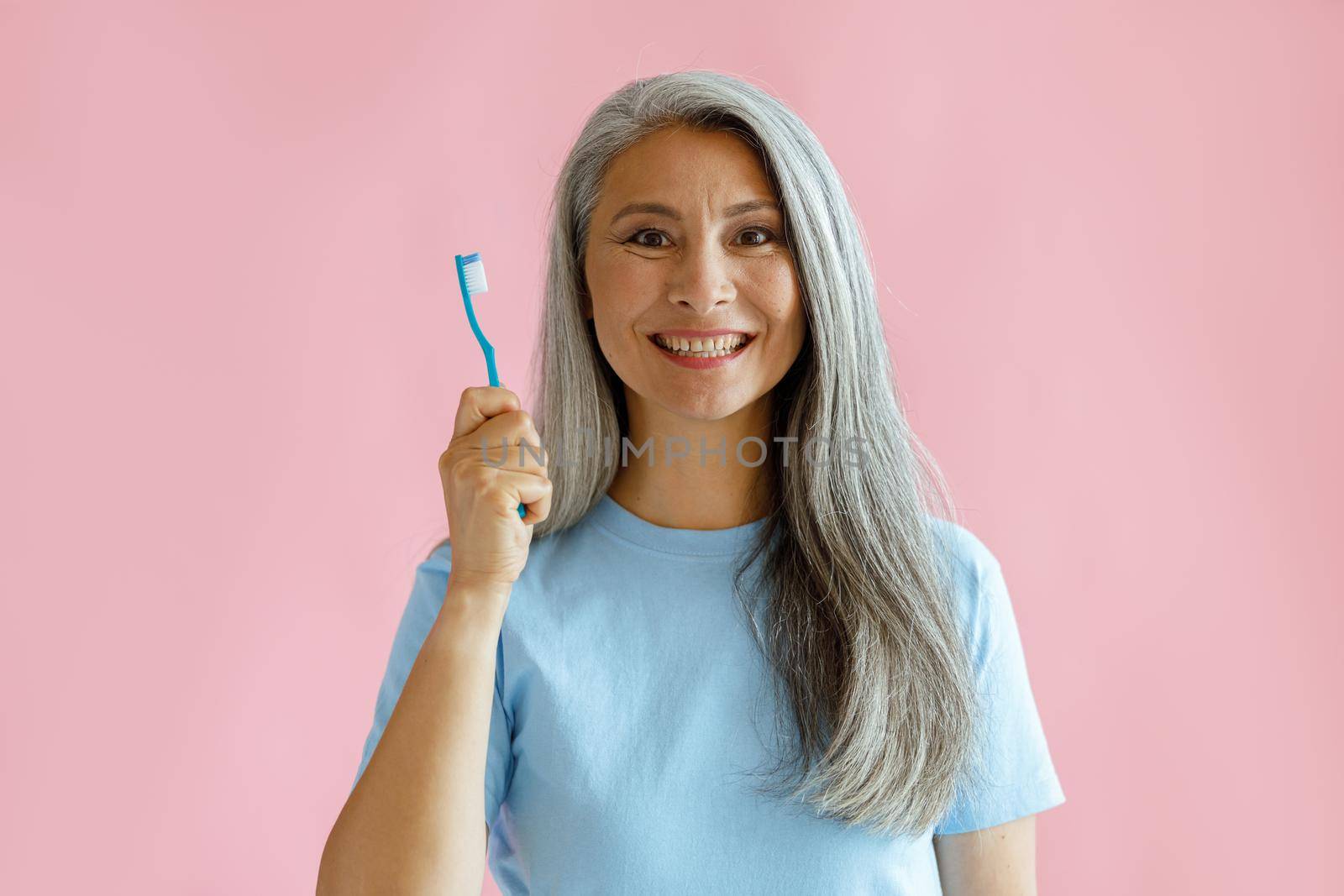 Cheerful middle aged Asian woman with grey hair in blue t-shirt holds toothbrush standing on pink background in studio. Oral cavity hygiene