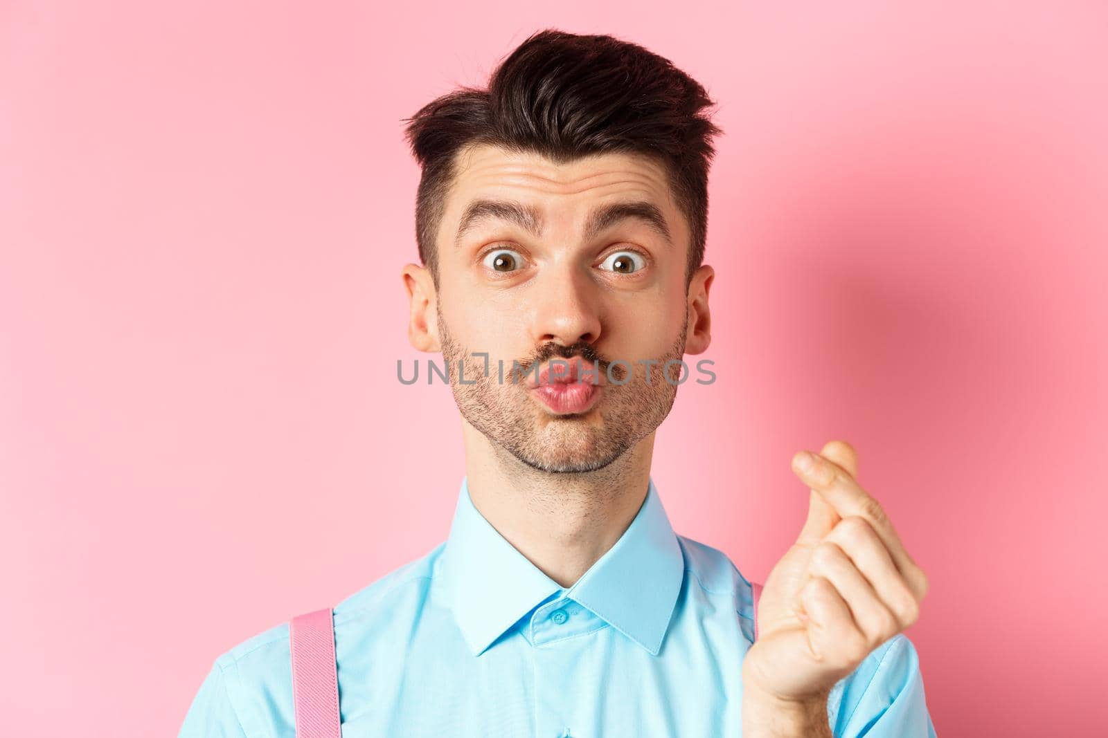 Valentines day concept. Silly guy waiting for kiss and showing finger heart gesture, express love and sympathy, pink background.