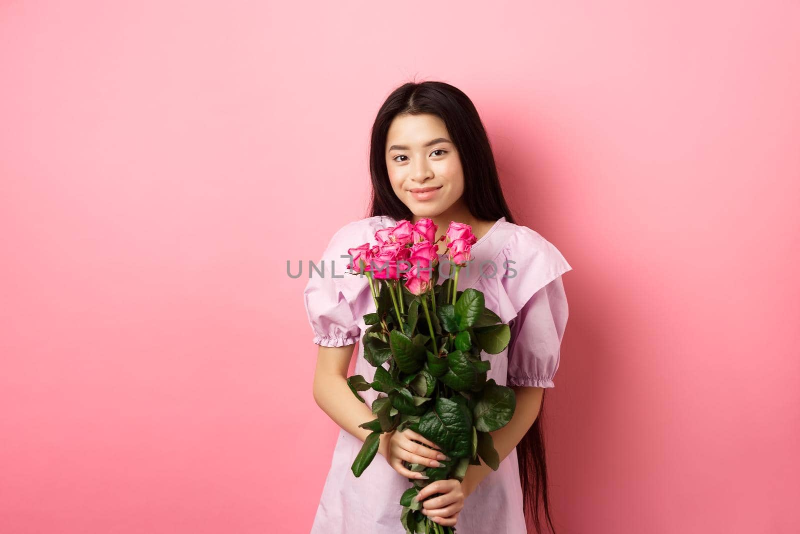 Romantic tender asian girl holding bouquet of roses, smiling cute at camera, having valentines date with lover, wearing dress, pink background by Benzoix
