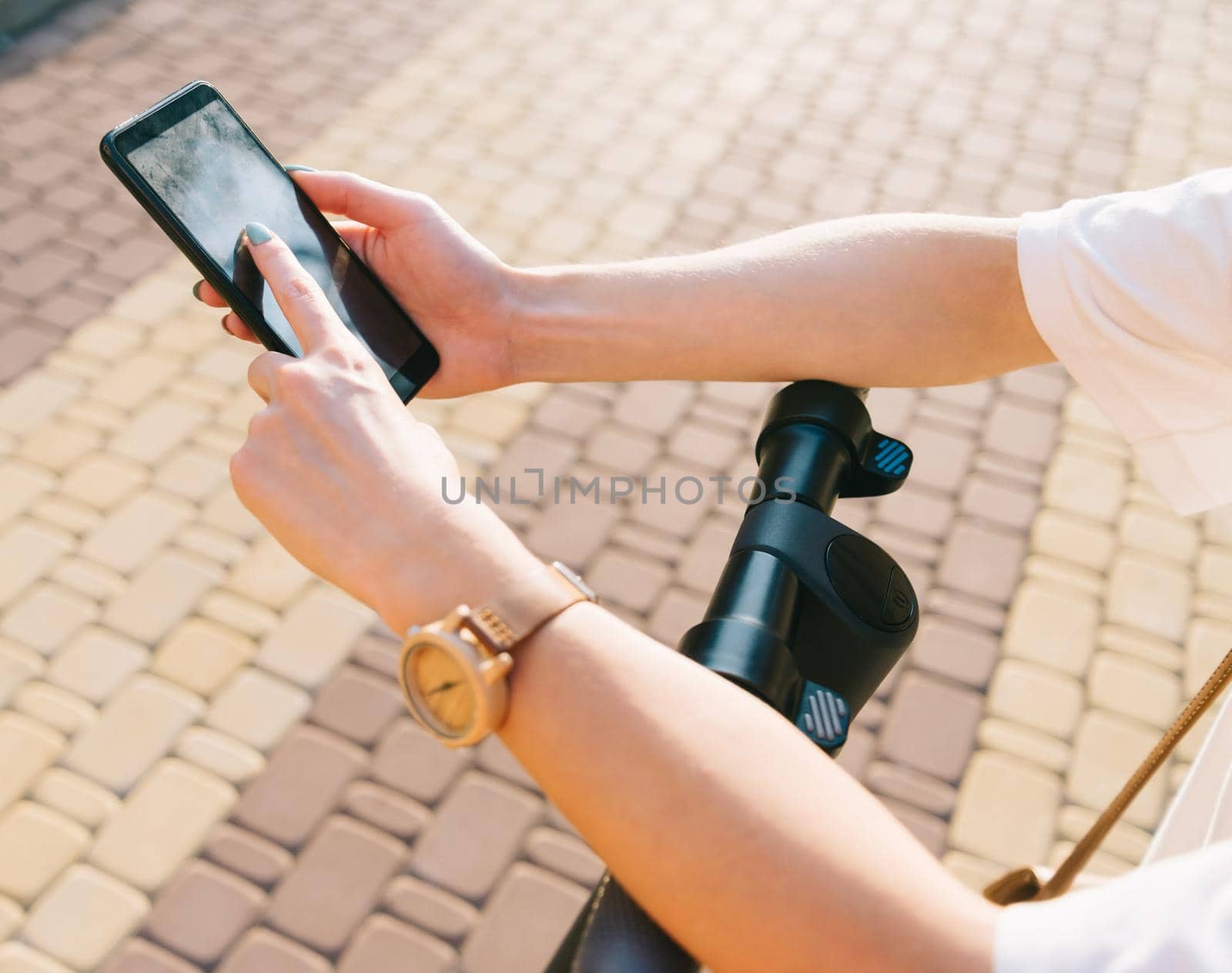 View of female hands typing text message and using smartphone while standing with an electric scooter in city street.