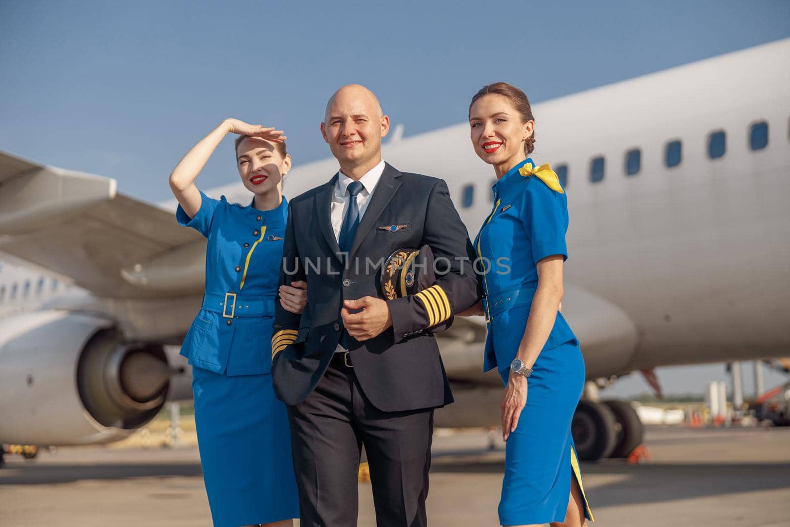Happy pilot and two attractive stewardesses standing together in front of an airplane and smiling after landing. Aircraft, aircrew, occupation concept