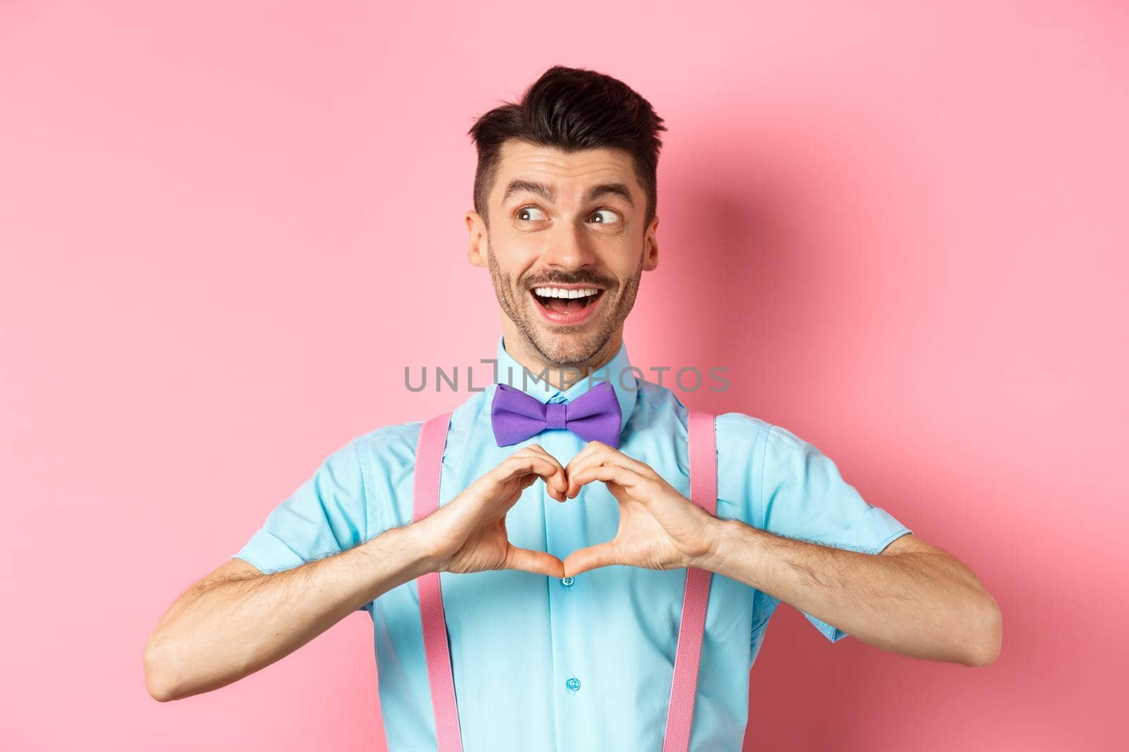 Happy Valentines day. Cheerful male model enjoying romantic date, showing heart sign, say I love you and looking left with happy smile, standing over pink background by Benzoix