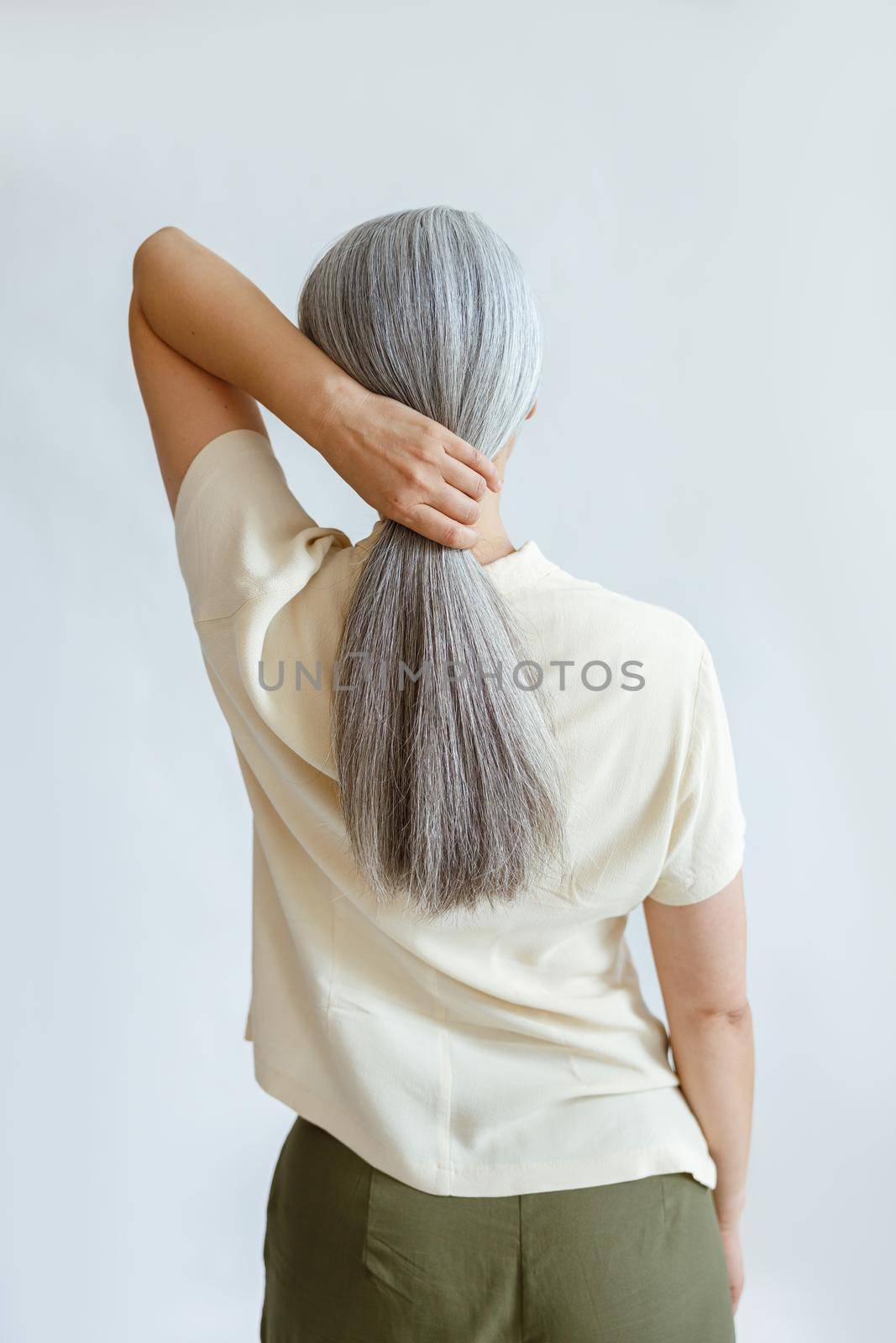 Elegant woman in yellow shirt makes polytail of long silver hair standing on light grey background in studio backside view. Mature beauty lifestyle