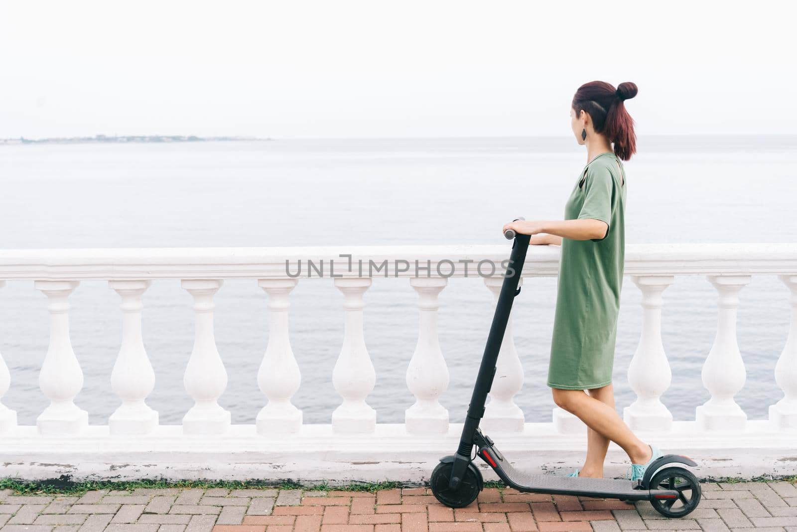 Modern girl in dress standing with an electric scooter on embankment and looking at the sea in summer.