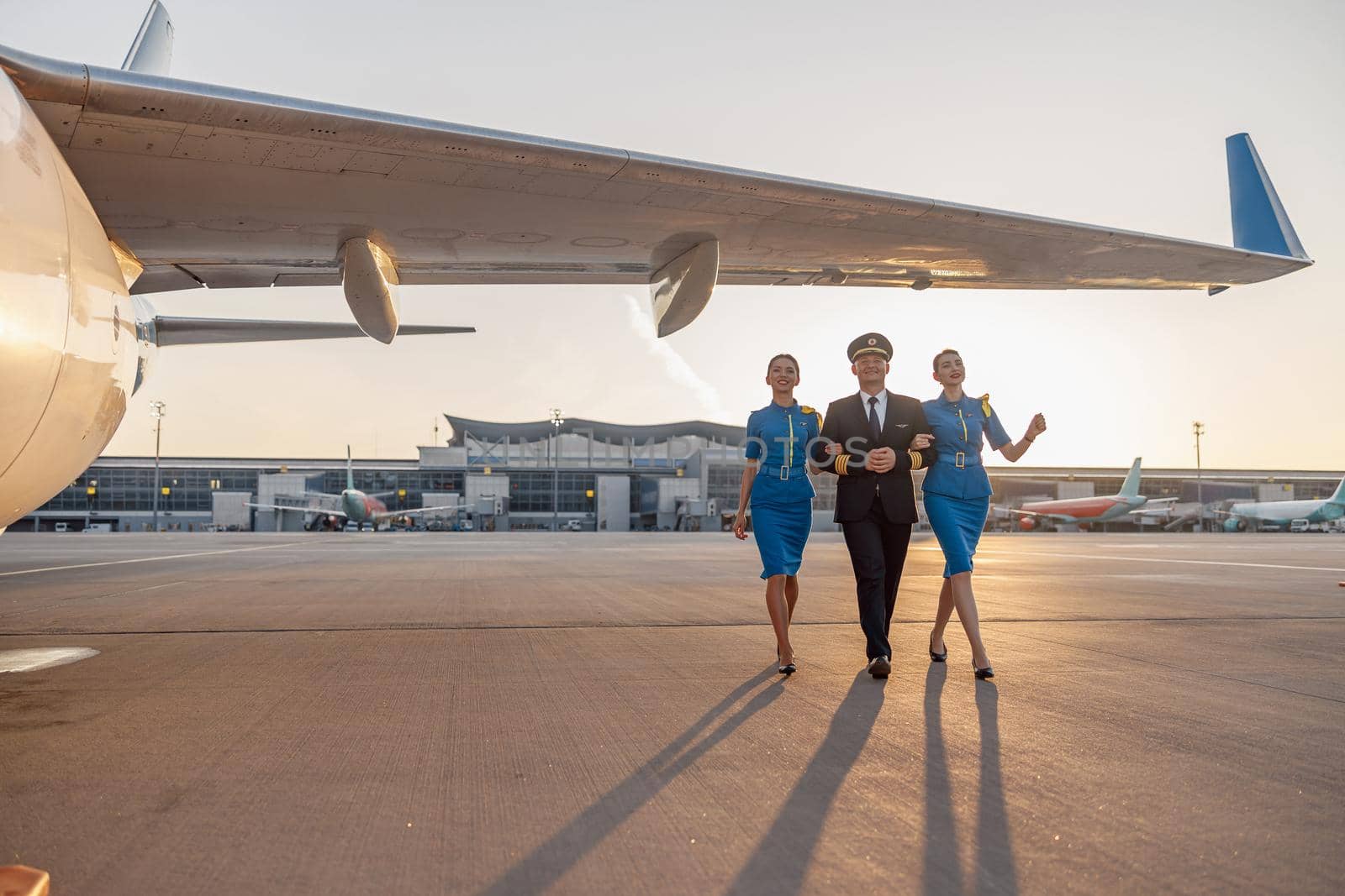 Full length shot of excited male pilot walking together with two female flight attendants in blue uniform in front of an airplane in terminal at sunset. Aircraft, aircrew concept