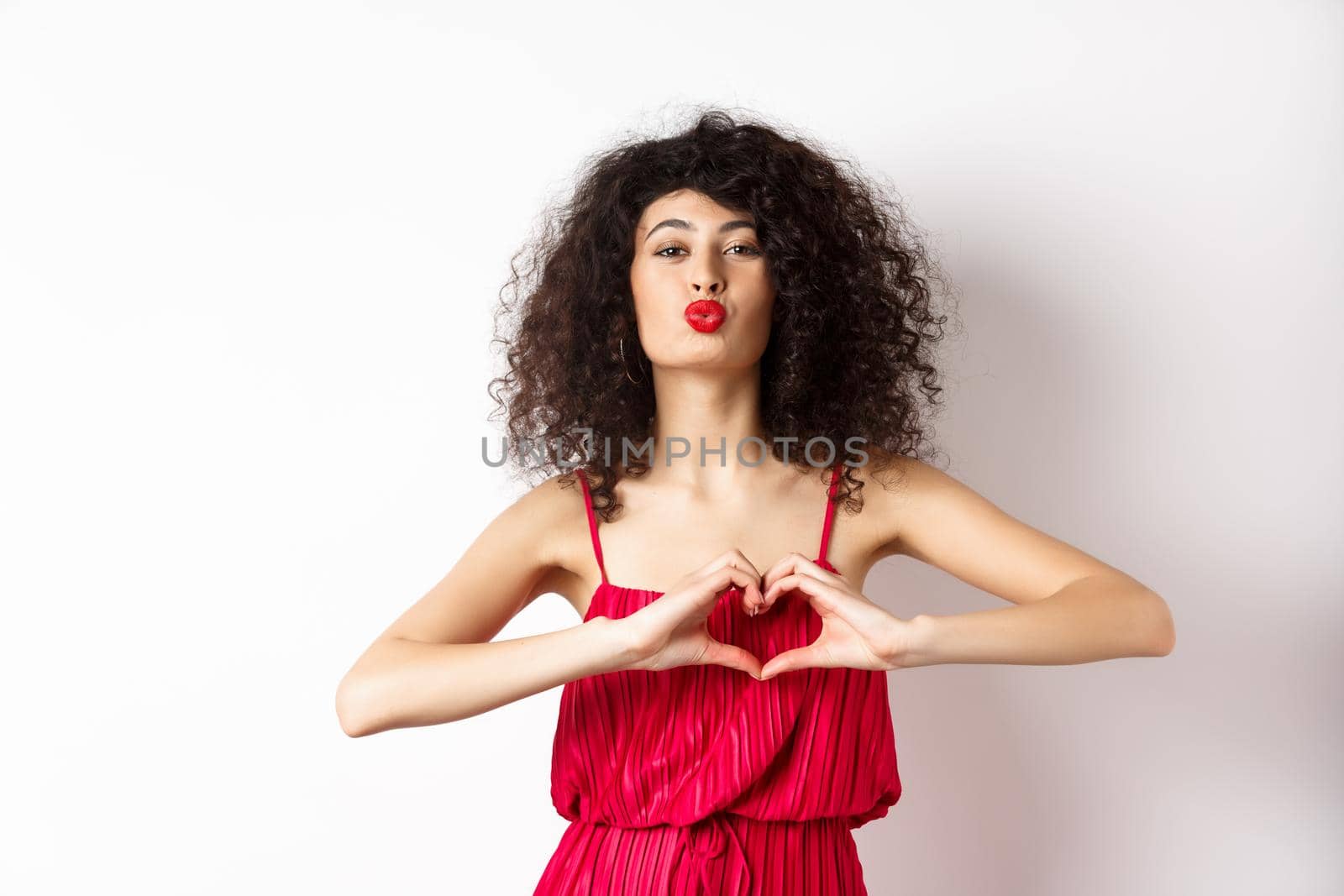 Beautiful lady with curly hair, red dress, showing heart symbol and pucker lips for kiss, love you gesture, standing on white background.