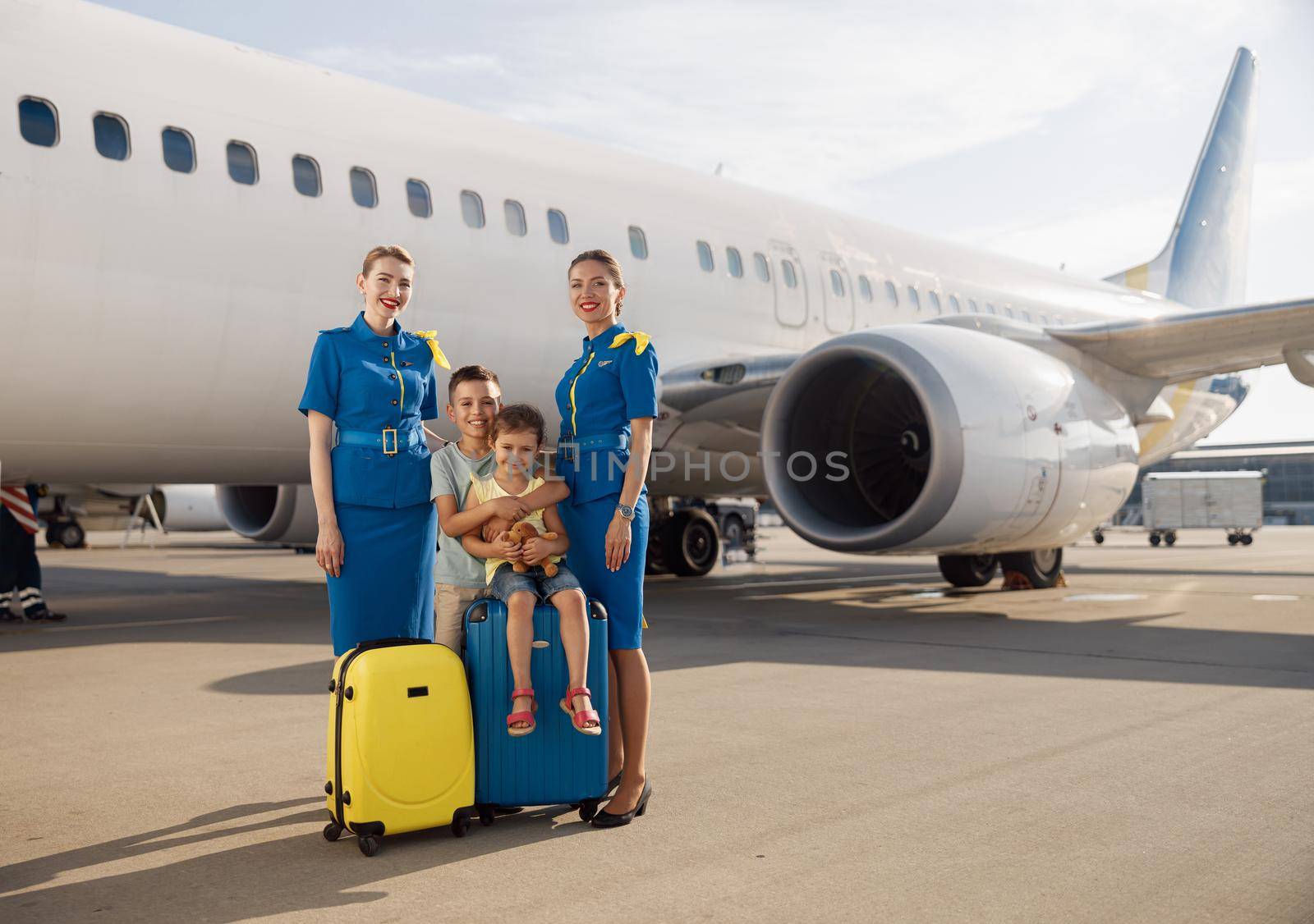Full length shot of happy two kids sitting on their luggage in front of big airplane on a daytime and smiling at camera together with beautiful stewardesses by Yaroslav_astakhov