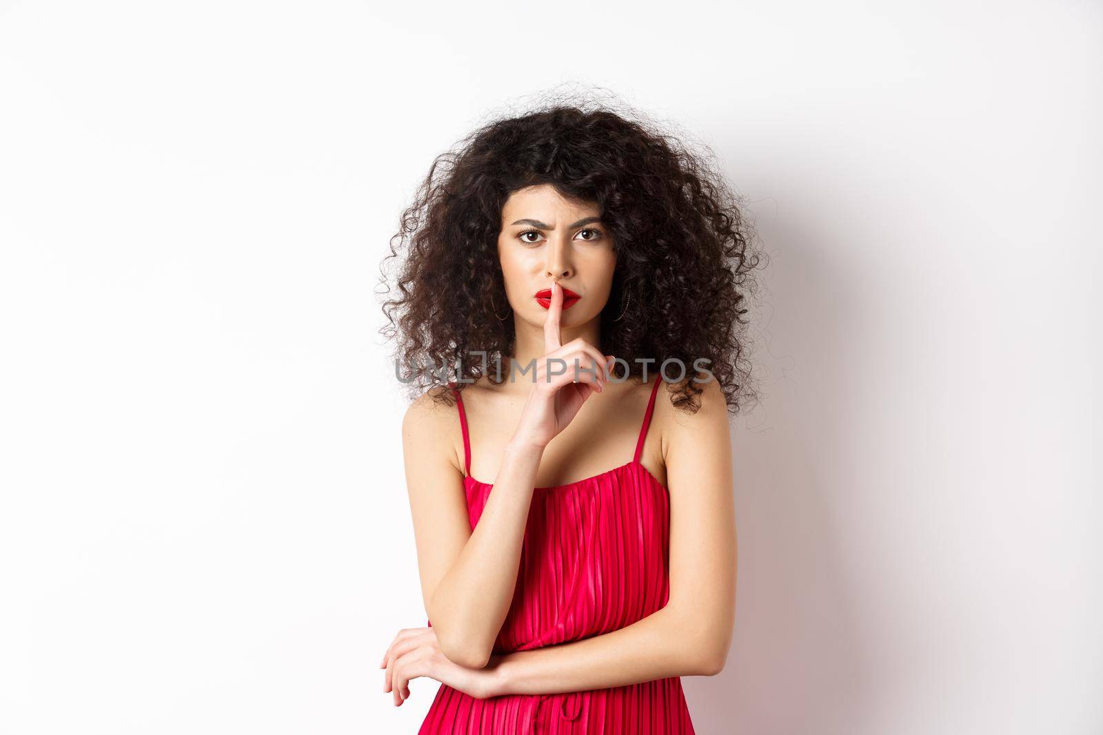 Angry elegant woman in red dress hushing and frowning, tell to be quiet, asking for silence, standing over white background.