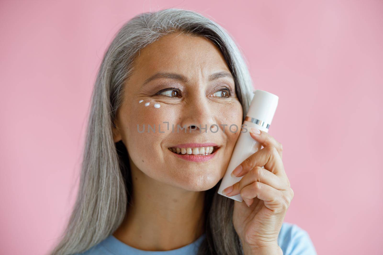 Positive middle aged Asian model with cream drops under eye holds bottle on pink background in studio. Mature beauty lifestyle