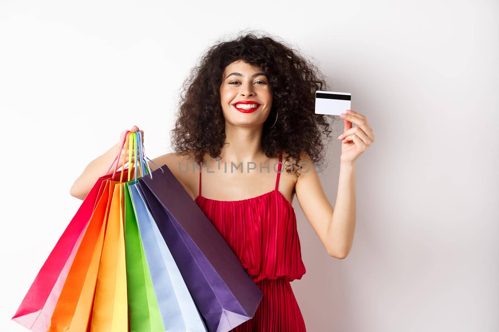 Happy elegant woman in red dress, showing shopping bags and plastic credit card, smiling pleased, standing on white background by Benzoix