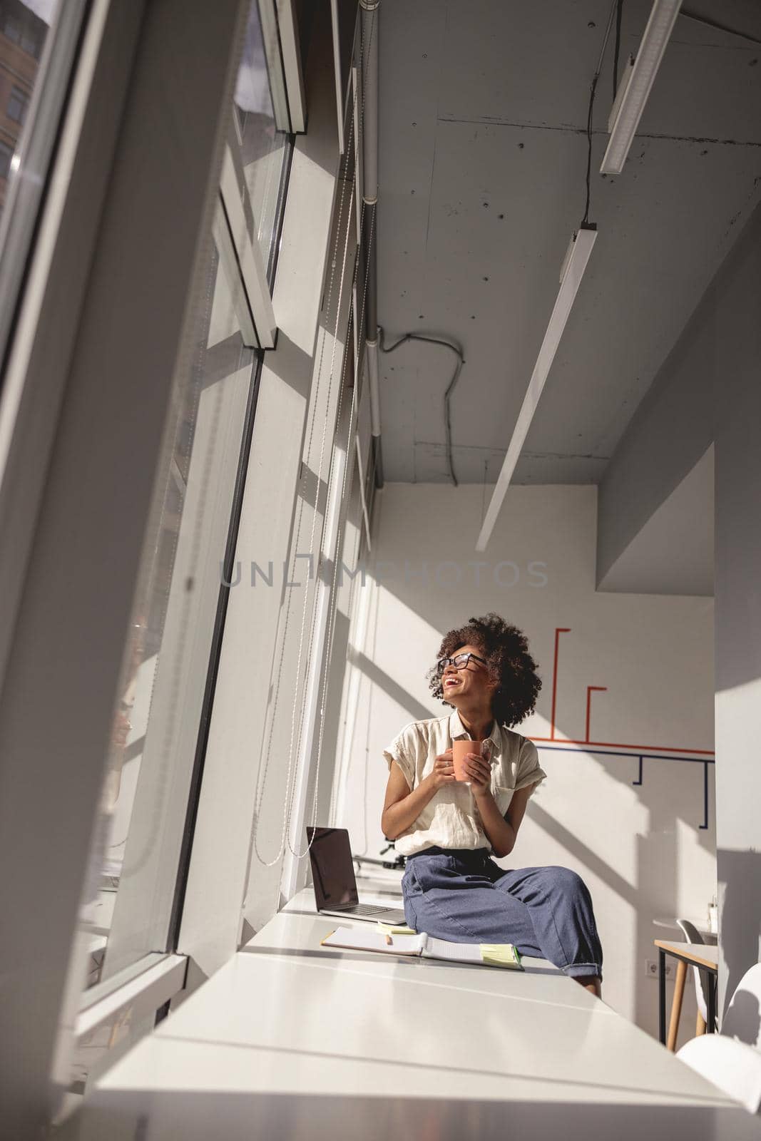 Happy Afro American woman enjoying sunny day in the office while holding cup of coffee