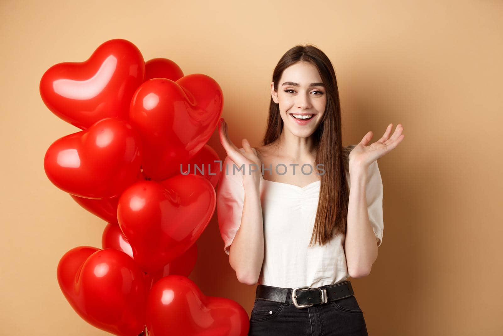 Valentines day and love concept. Happy smiling woman raising hands up surprised, standing near romantic heart balloons on beige background.