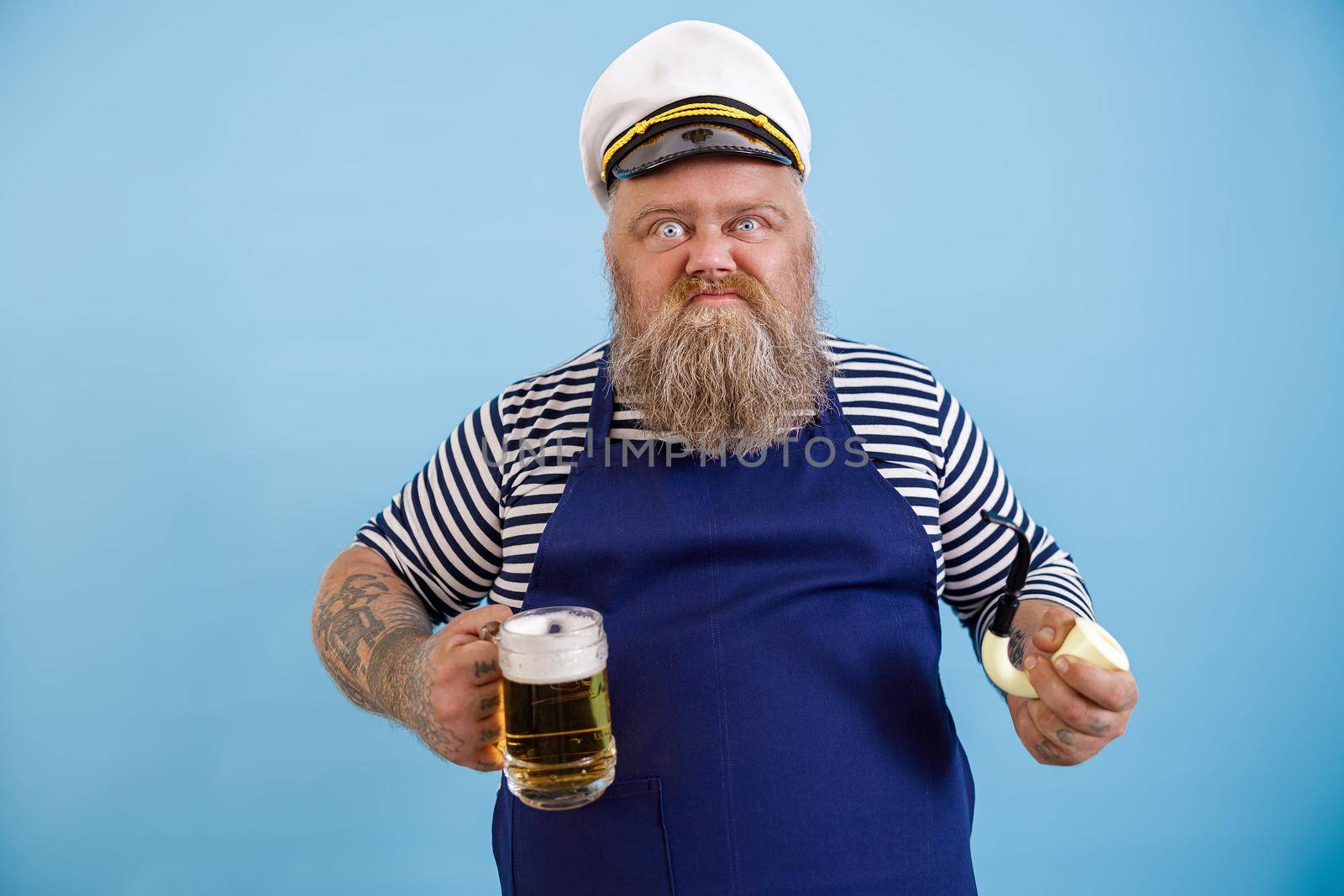 Funny aggressive obese man in sailor costume with apron holds smoking pipe and glass mug of delicious beer on light blue background in studio