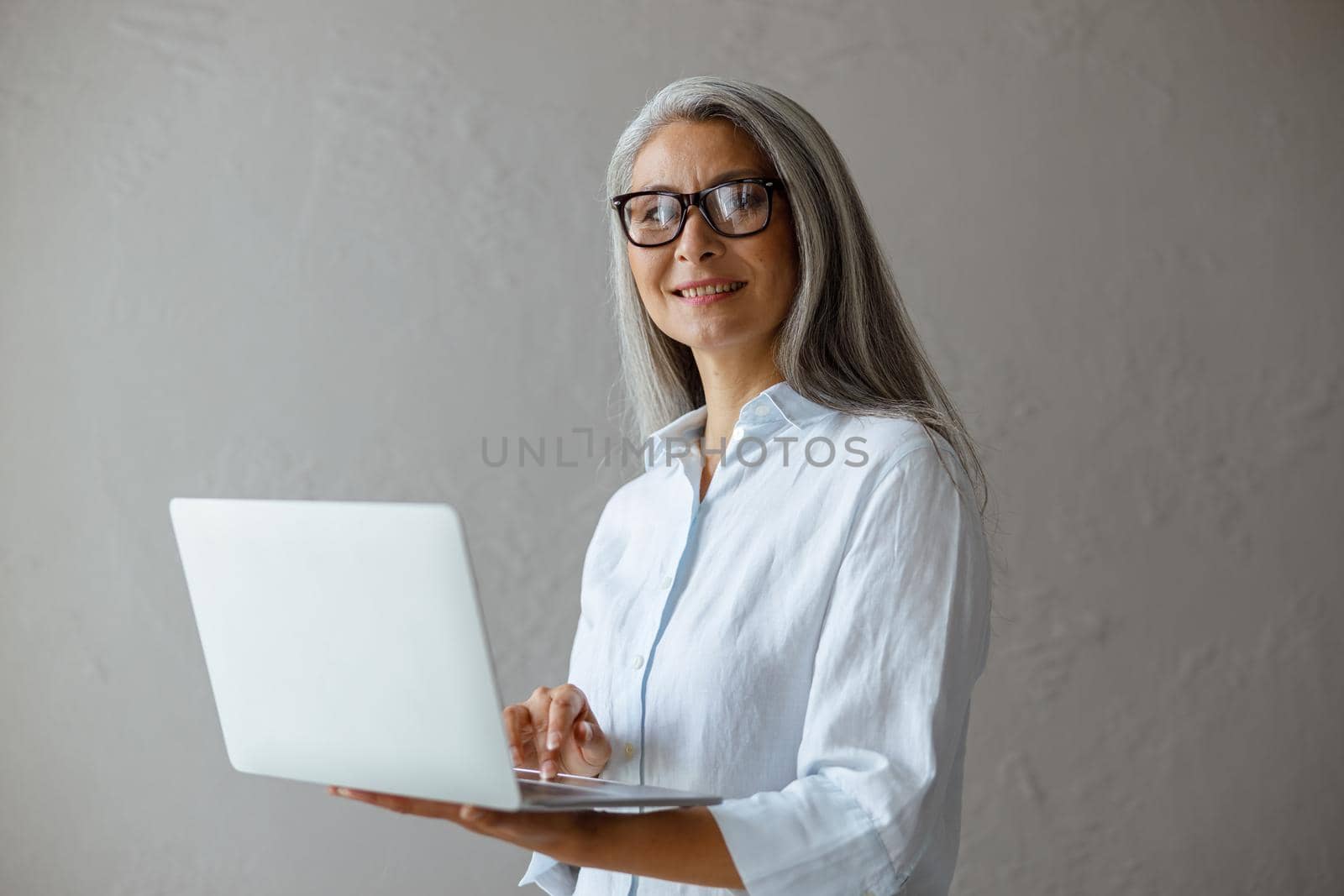Smiling silver haired middle aged Asian woman in white blouse works on modern laptop standing near grey stone wall in studio