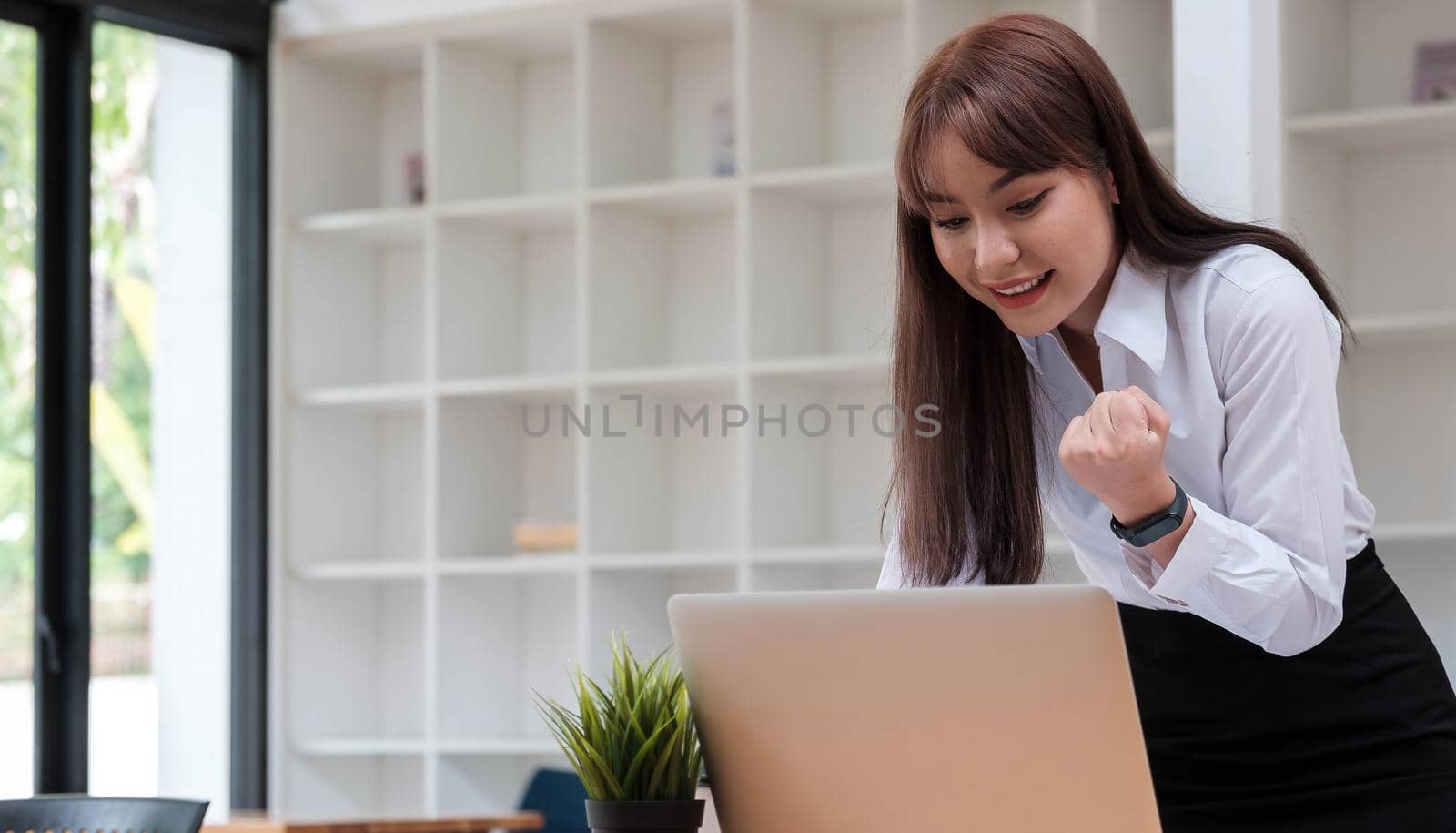 Photo of a cheerful business woman standing in a conference room with a laptop computer. looking aside make winner gesture.