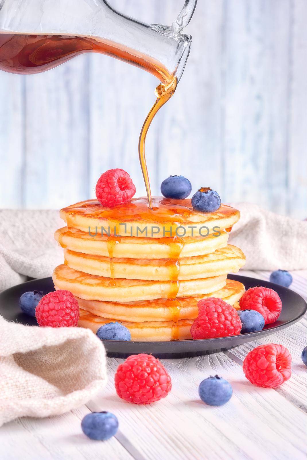 Delicious pancakes with fresh blueberries, raspberries are poured with sweet maple syrup on a light wooden background.