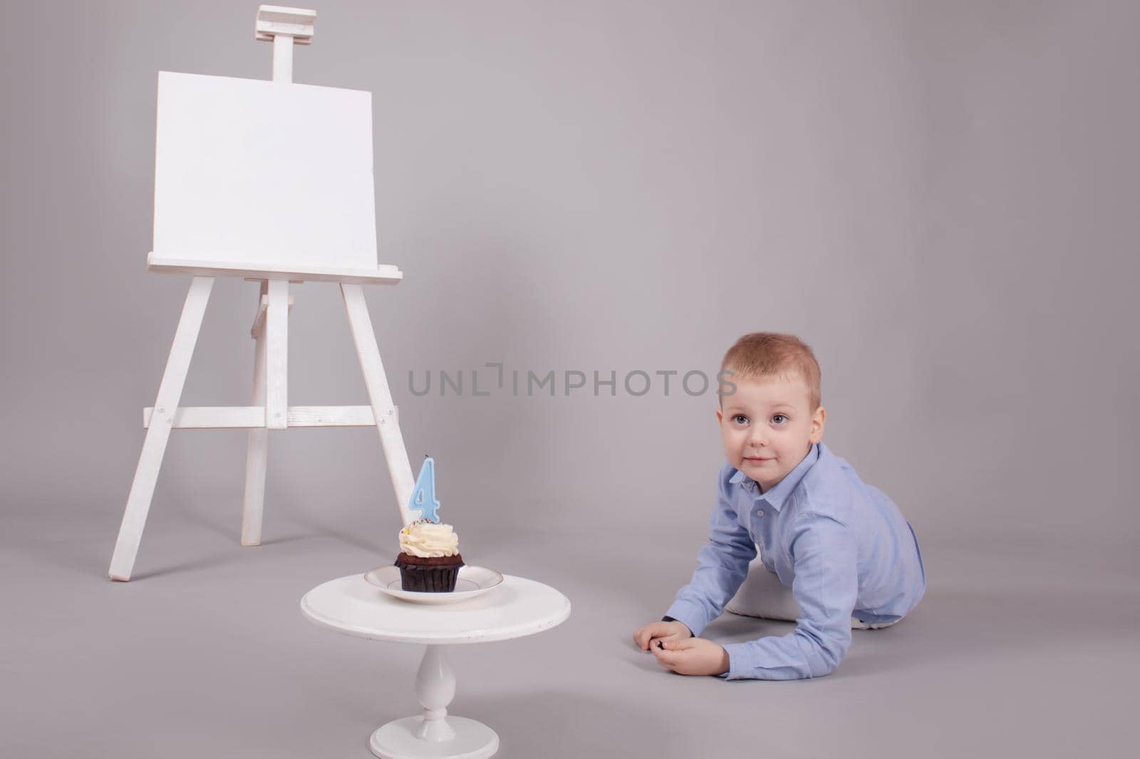 preschool boy in white pants and blue shirt on grey background near easel with mockup and cupcake with candle, shaped in number four 4. happy birthday. celebration. High quality photo