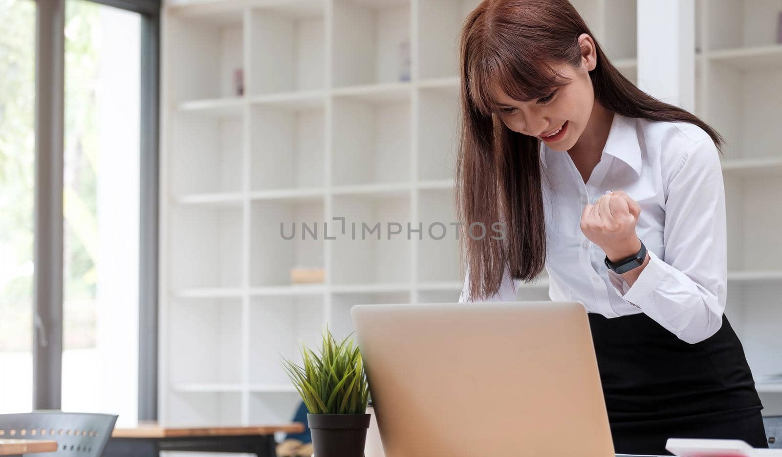 Photo of a cheerful business woman standing in a conference room with a laptop computer. looking aside make winner gesture.